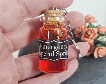 May include: A small glass bottle keychain with a cork stopper filled with a red liquid and gold glitter. The bottle has a black label that reads "Emergency Aperol Spritz".