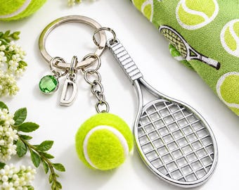May include: A silver-toned metal keychain featuring a tennis racket charm, a miniature fuzzy yellow tennis ball, a small green crystal bead, and a silver letter D charm attached to a circular split ring.