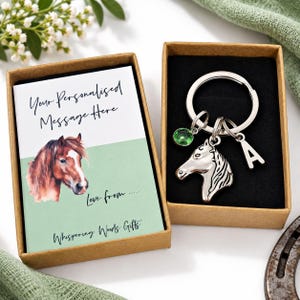 Op de afbeelding: Een zilverkleurige sleutelhanger met een paardenkopbedel, een letter A-bedel en een ronde groene kristallen bedel, gepresenteerd in een bruine kartonnen geschenkdoos met een kaart met een paardenillustratie en de tekst Your Personalized Message Here.
