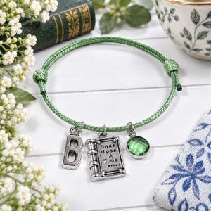 May include: A green cord bracelet featuring three silver-toned charms: a letter B, a rectangular book-shaped charm with the text Once Upon A Time, and a round, faceted green crystal bead. The bracelet has adjustable sliding knots.
