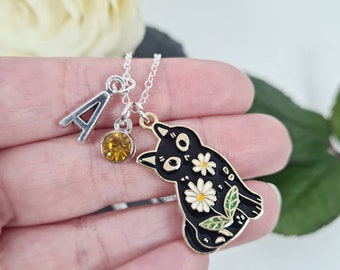May include: A silver chain necklace with a black cat charm. The cat charm has white flowers and green leaves. The necklace also has a silver charm with the letter A and a yellow crystal.