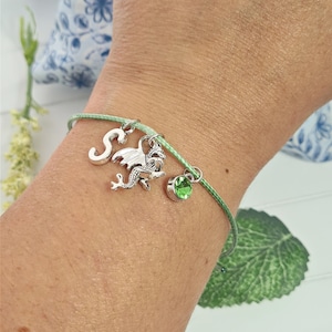May include: A green string bracelet with a silver charm of a dragon, a silver charm with the letter 'S', and a green gemstone charm.