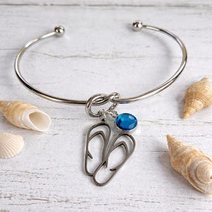Op de afbeelding: Een zilverkleurige armband met een knoopontwerp en een bedel met slippers en een blauwe edelsteen. De armband ligt op een wit houten oppervlak, met schelpen eromheen. Een stijlvol accessoire.