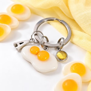 May include: A silver-toned metal keyring featuring a charm shaped like a fried egg with two yellow yolks, a silver letter A charm, and a small round yellow faceted crystal charm attached to a circular split ring.