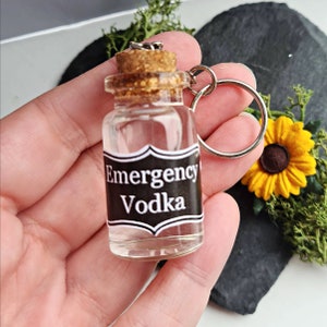 May include: A small glass bottle with a cork top and a black label that reads "Emergency Vodka". The bottle is attached to a silver key chain.