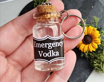 May include: A small glass bottle with a cork top and a black label that reads "Emergency Vodka". The bottle is attached to a silver key chain.