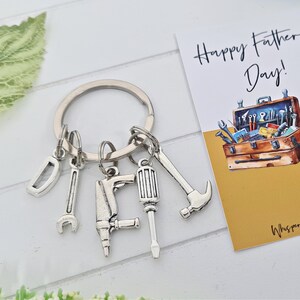 May include: A silver keyring with a variety of tool charms, including a wrench, screwdriver, hammer, and drill. The keyring is attached to a silver key ring. The keyring is accompanied by a card that reads "Happy Father's Day!" with a watercolour illustration of a toolbox filled with tools.