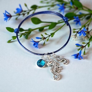 Op de afbeelding: Een donkerblauwe armband met een zeepaardjesbedel, een blauwe edelsteen en de letter "S". De armband is versierd met kleine blauwe bloemen en groene bladeren. De armband ligt op een witte ondergrond.