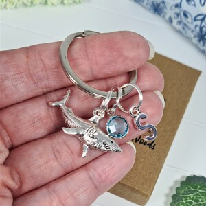 Puede incluir: Llavero de tibur&oacute;n plateado con un dije de piedra preciosa azul y un dije de letra 'S' plateado. El llavero est&aacute; unido a un anillo plateado.
