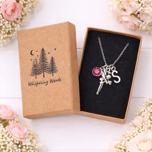 May include: A silver necklace with a fairy charm, a pink gemstone, and the letter "S" is presented in an open gift box. The box is brown with the words "Whispering Woods" and a tree design. The necklace is on a black background.