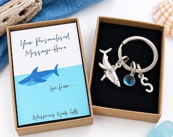 May include: A silver-toned shark keychain with a circular ring, a small blue circular charm, and a silver letter S charm. The keychain is presented in a brown cardboard box alongside a card featuring a blue shark illustration and text.