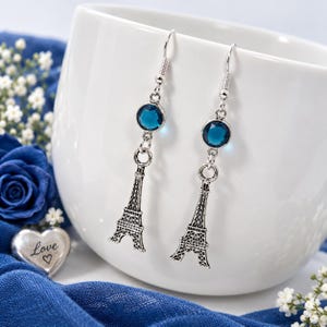 May include: A pair of silver-toned drop earrings featuring a round, dark blue faceted crystal bead positioned above a detailed, miniature metal Eiffel Tower charm. The earrings are suspended from standard silver-toned metal fishhook ear wires.