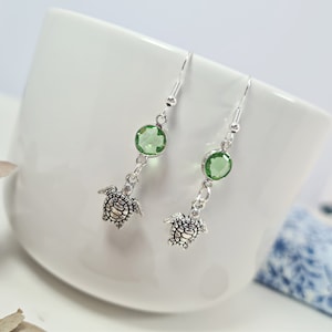 May include: Silver-coloured drop earrings with small turtle charms and round, light green gemstones. The earrings have a hook closure and are displayed against a white background. The earrings are a jewelry item.