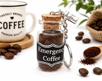 May include: A clear glass bottle keychain filled with dark brown coffee grounds, featuring a cork stopper and a black label with white text that reads Emergency Coffee. A small silver charm attached to the keychain ring has the letters fee visible.