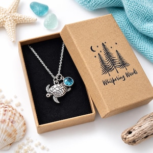 May include: A silver-toned necklace featuring a sea turtle pendant and a round blue crystal charm, presented in a brown cardboard gift box with a black insert. The box lid is printed with a black pine tree graphic and the text Whispering Woods.