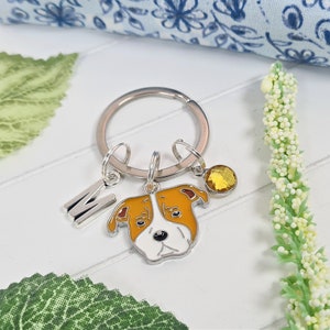 May include: A silver key ring with a charm of a brown and white bulldog, a silver letter M charm, and a yellow crystal charm.