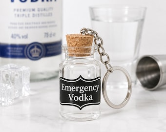 Może przedstawiać: Brelok w kształcie miniaturowej butelki z przezroczystego szkła z korkiem i czarną etykietą z białym napisem Emergency Vodka. Butelka jest przymocowana do srebrnego metalowego łańcuszka i okrągłego kółka na klucze.