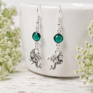 May include: Silver-toned dangle earrings featuring a round, emerald-green gemstone and a detailed dragon charm. The earrings have a fish hook closure. The background is a white ceramic container and white flowers.