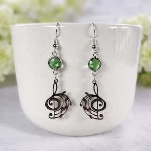Op de afbeelding: Een paar zilverkleurige oorbellen met groene edelsteenaccenten en muzieknootbedels. De oorbellen hebben een ronde groene edelsteen, een zilveren ketting en een donkerbruin G-sleutelontwerp met muzieknoten.