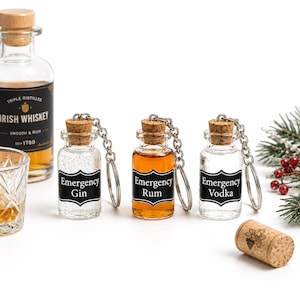 May include: Three miniature glass bottle keychains with cork stoppers and silver-tone metal chains. Each bottle features a black label with white text reading Emergency Gin, Emergency Rum, or Emergency Vodka. The bottles contain clear or amber liquids.