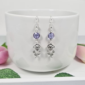 May include: A pair of silver earrings featuring a delicate flower design and a purple crystal bead. The earrings are hanging against a white background.