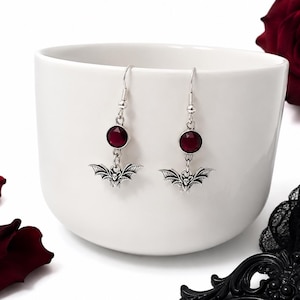 May include: A pair of silver-toned drop earrings featuring a round, faceted deep red gemstone set above a small, detailed metal bat charm. The earrings hang from standard silver-toned ear wires, showcasing a symmetrical design with dark accents.