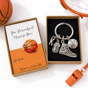 May include: A silver-toned metal keyring featuring a letter K charm, a baseball cap charm, a high-top sneaker charm, and a basketball charm, presented in a brown cardboard gift box with a card reading Your Personalised Message Here.