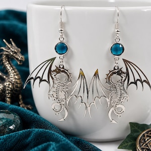 May include: A pair of silver-toned dangle earrings featuring a detailed dragon silhouette with outstretched wings. Each earring is accented with a single round, faceted dark blue crystal bead positioned above the dragon charm on a silver hook.