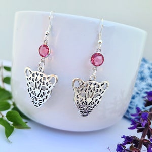 May include: Silver earrings featuring a pink crystal and a detailed cutout of a leopard's face.