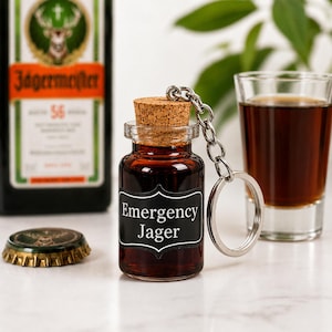 May include: A small clear glass bottle keyring filled with dark brown liquid, topped with a cork stopper and a silver metal chain. The bottle features a black label with white text that reads Emergency Jager in a decorative frame.