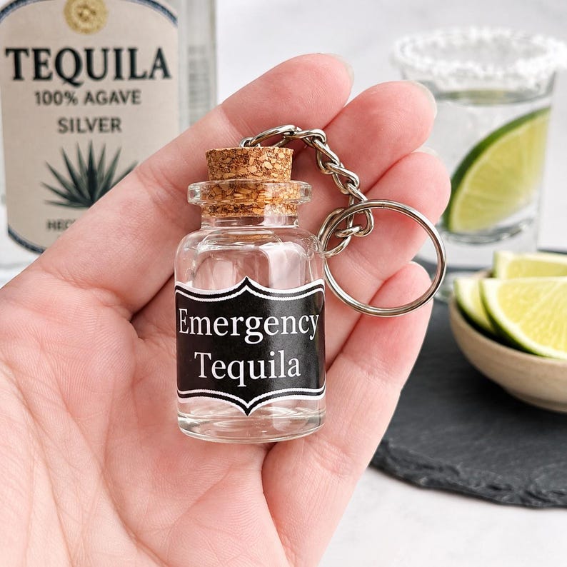 Peut inclure: Un porte-cl&eacute;s en forme de bouteille miniature en verre transparent avec un bouchon en li&egrave;ge et une &eacute;tiquette noire portant le texte blanc Emergency Tequila. La bouteille est fix&eacute;e &agrave; une cha&icirc;ne en m&eacute;tal argent&eacute; et un anneau bris&eacute;.