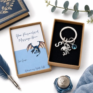 May include: A silver-toned metal keyring featuring a dragon charm, a letter S charm, and a round blue crystal charm, presented in a brown cardboard box with a card that reads Your Personalised Message Here, Love from, and Whispering Woods Gifts.