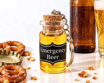 May include: A clear glass miniature bottle keyring filled with a golden liquid, topped with a cork stopper. A black label on the front features the white text Emergency Beer. The keyring includes a silver metal ring and chain attachment.