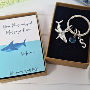 Puede incluir: Una peque&ntilde;a caja de regalo con una ilustraci&oacute;n de tibur&oacute;n azul y blanco y el texto "Your Personalised Message Here" y "Love from..." La caja est&aacute; abierta y muestra un llavero de tibur&oacute;n plateado con un dije de cristal azul y un dije de letra "S" plateado.