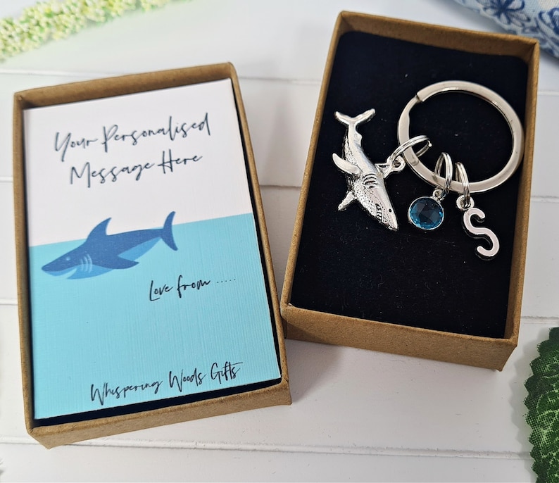 Puede incluir: Una peque&ntilde;a caja de regalo con una ilustraci&oacute;n de tibur&oacute;n azul y blanco y el texto "Your Personalised Message Here" y "Love from..." La caja est&aacute; abierta y muestra un llavero de tibur&oacute;n plateado con un dije de cristal azul y un dije de letra "S" plateado.