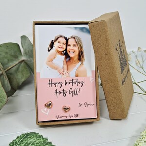 May include: A pink card with the text "Happy birthday, Aunty Gill! Love Sophie x" and a photo of two women. The card is in a brown box with a small tree design. The card is accompanied by a pair of gold heart-shaped earrings.