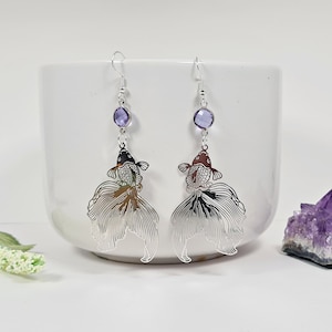 May include: Silver-toned dangle earrings featuring a fish design. Each earring has a purple gemstone and a detailed fish silhouette with a flowing tail. The fish heads are black and brown.