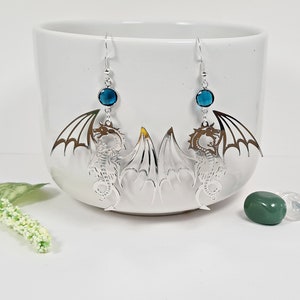 May include: Silver dragon earrings with blue crystal accents. The dragon is a detailed silhouette with wings outstretched.