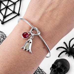 Könnte beinhalten: Ein silberfarbenes Armband mit Knotendetail und zwei hängenden Anhängern: eine kleine, glatte silberfarbene Geisterfigur mit zwei Augenausschnitten und ein runder, facettierter, tiefroter Edelstein in einer silberfarbenen Metallfassung.