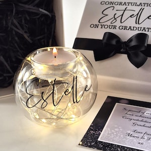 May include: A glass candle holder with the name "Estelle" in script, illuminated by a candle and fairy lights. Beside it is a white gift box with a black ribbon and the words "Congratulations Estelle on your Graduation".