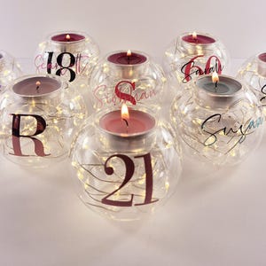 May include: A collection of eight spherical clear glass candle holders, each containing thin copper wire fairy lights and a lit tea light candle. The glass surfaces feature various names and numbers in black and pink script and block lettering.