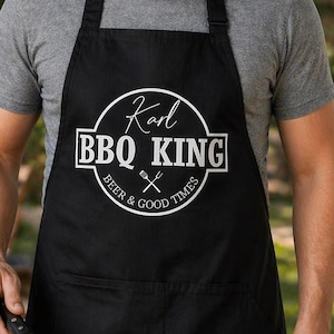 Pu&ograve; includere: Grembiule nero con la scritta "Karl BBQ KING" in un design circolare bianco. Il grembiule ha anche le parole "Beer & Good Times" e una forchetta e un coltello incrociati. La persona sta grigliando cibo su un barbecue.