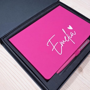 May include: A vibrant pink notebook and pen set, presented in a black gift box. The notebook is inscribed with the name "Emelia" in white script, accompanied by a heart. The pen matches the notebook's colour.