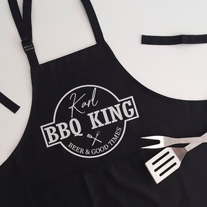 Pu&ograve; includere: Grembiule nero con la scritta bianca "Karl BBQ KING BEER & GOOD TIMES". Il grembiule ha un cinturino regolabile per il collo e lacci in vita. Una spatola e una forchetta sono a destra.