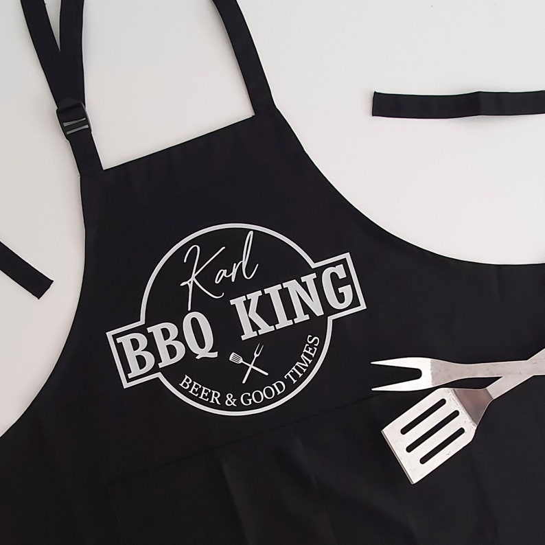 Pu&ograve; includere: Grembiule nero con la scritta bianca "Karl BBQ KING BEER & GOOD TIMES". Il grembiule ha un cinturino regolabile per il collo e lacci in vita. Una spatola e una forchetta sono a destra.