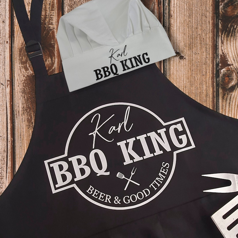 Pu&ograve; includere: Un grembiule nero e un cappello da chef bianco con la scritta "Karl BBQ KING". Il grembiule presenta un design circolare con lo stesso testo, pi&ugrave; "Beer & Good Times" e una grafica di forchetta e spatola. Il cappello &egrave; bianco con un bordo nero.
