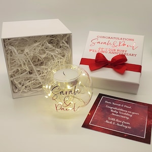 May include: A white gift box with a red ribbon and the text "Congratulations Sarah & Paul on your Ruby Wedding Anniversary". A clear glass globe with a candle inside and the text "Sarah & Paul" in red lettering. A red card with the text "Dear Sarah & Paul, Congratulations on your Ruby Wedding Anniversary. With love from Mick & Audrey xx".