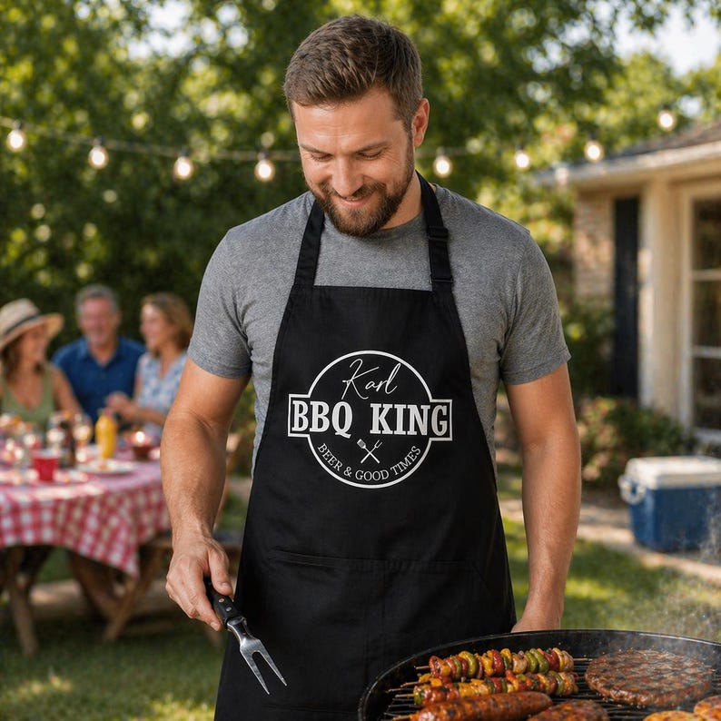 Pu&ograve; includere: Grembiule nero con la scritta "Karl BBQ KING" in un design circolare bianco. Il grembiule ha anche le parole "Beer & Good Times" e una forchetta e un coltello incrociati. La persona sta grigliando cibo su un barbecue.