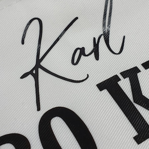 Pu&ograve; includere: Primo piano di un tessuto bianco con il nome "Karl" in nero in corsivo. Sotto il nome, sono parzialmente visibili grandi lettere nere. Il tessuto presenta un sottile motivo a trama diagonale.