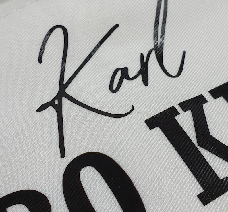Pu&ograve; includere: Primo piano di un tessuto bianco con il nome "Karl" in nero in corsivo. Sotto il nome, sono parzialmente visibili grandi lettere nere. Il tessuto presenta un sottile motivo a trama diagonale.